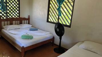 A room with a bed, a standing fan, and a window with a curtain.