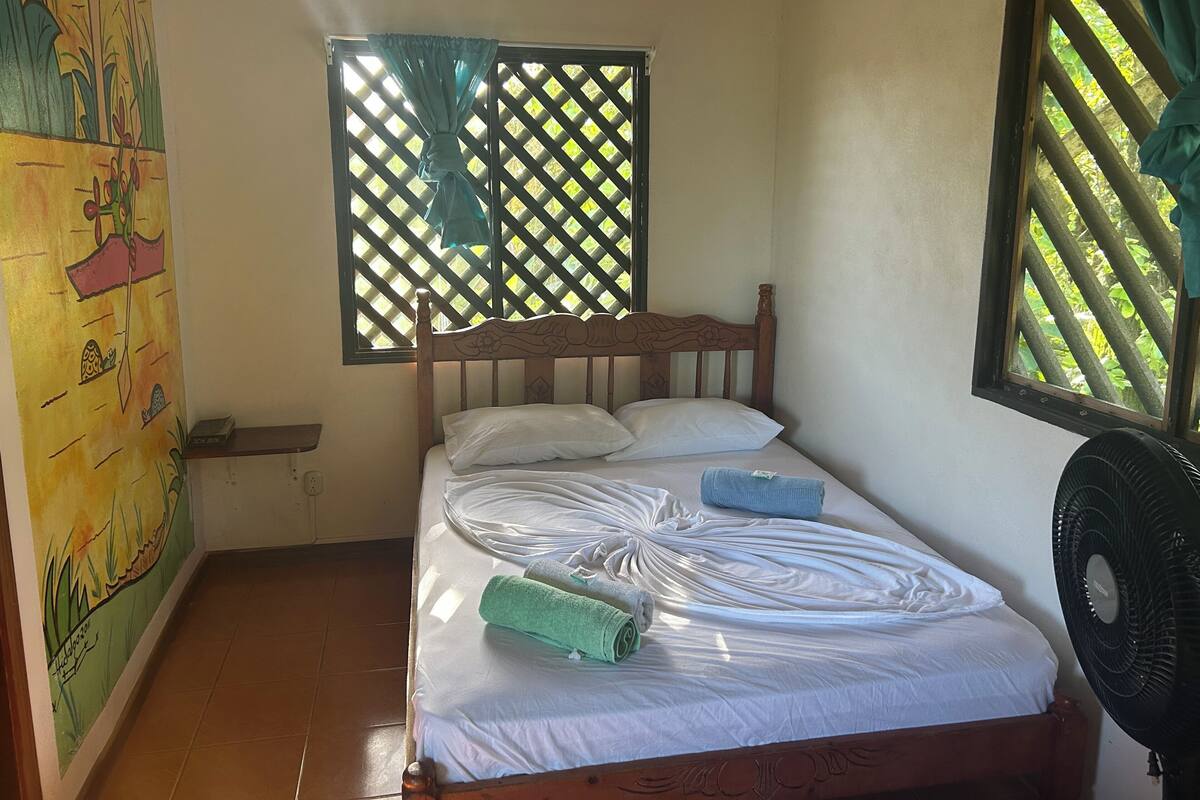 Ein Zimmer mit einem Bett, einem Fenster mit Gitterläden, einer Wand mit Wandgemälde und einem Standventilator.