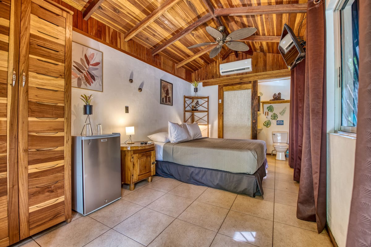 Une chambre à coucher comprenant un lit, une table de chevet, un réfrigérateur, un ventilateur de plafond et une fenêtre avec des rideaux.