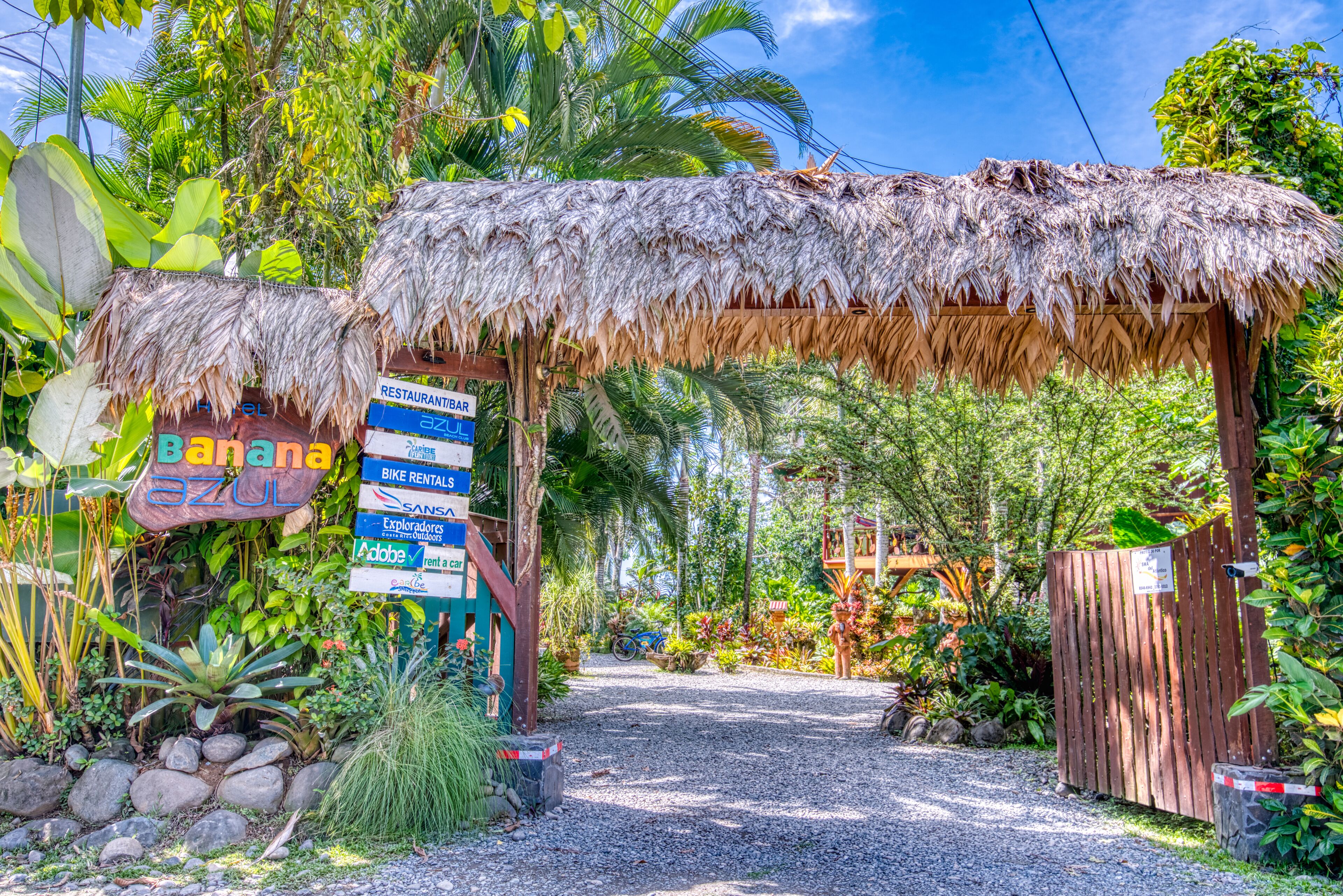 A tropical resort entrance with a thatched roof, a sign for 'Barana Azul' restaurant, and a bike rental sign.