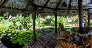 A wooden deck with a thatched roof, surrounded by lush greenery and a pond with lily pads.