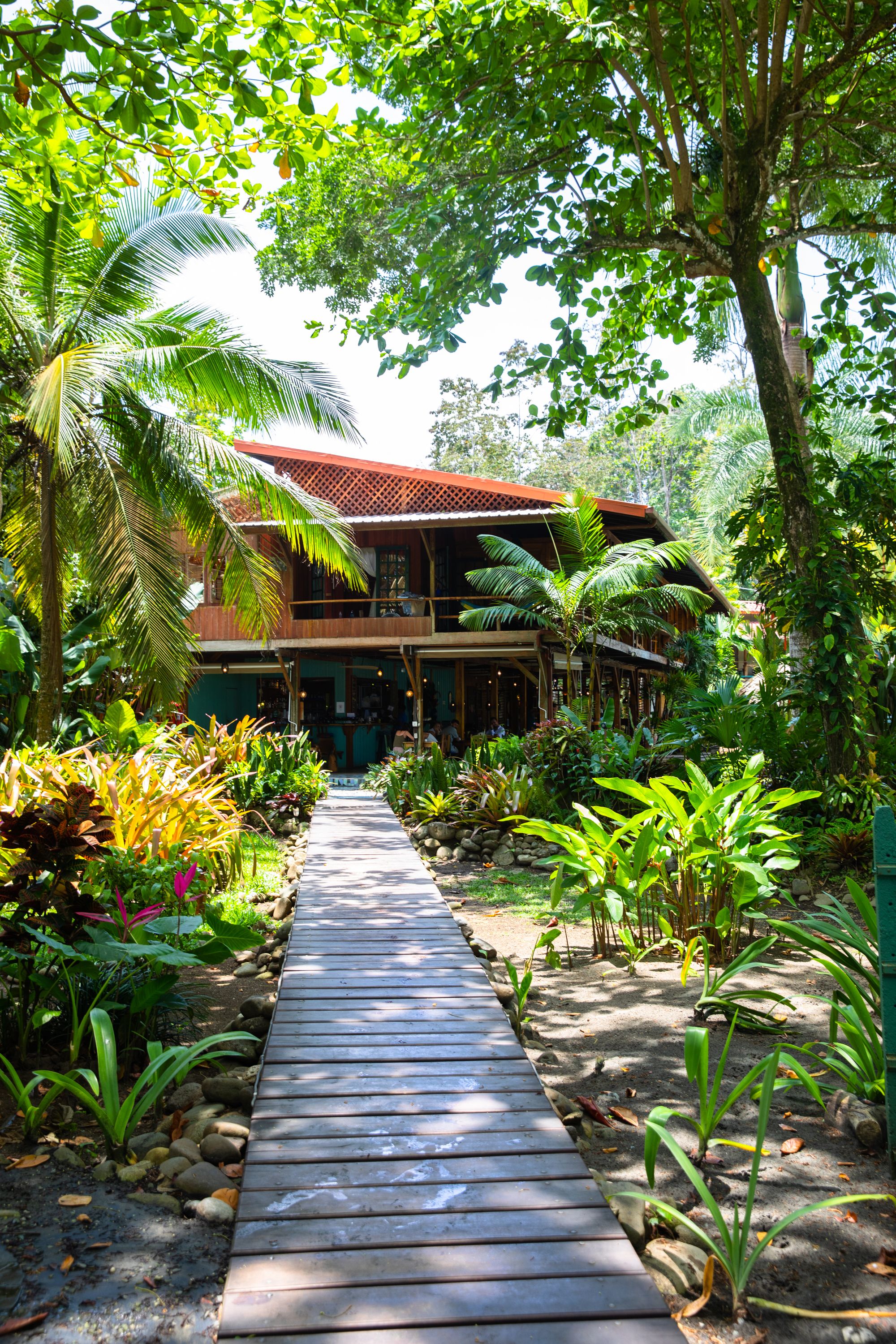 A wooden pathway leading to a building with a red roof, surrounded by lush greenery and palm trees.