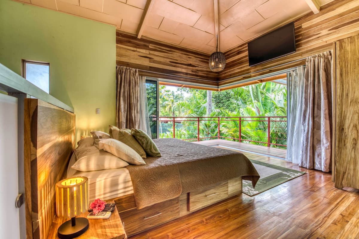 Une chambre dotée d’un grand lit, d’un plancher en bois, d’un téléviseur fixé au mur et d’un balcon offrant une vue sur la verdure.