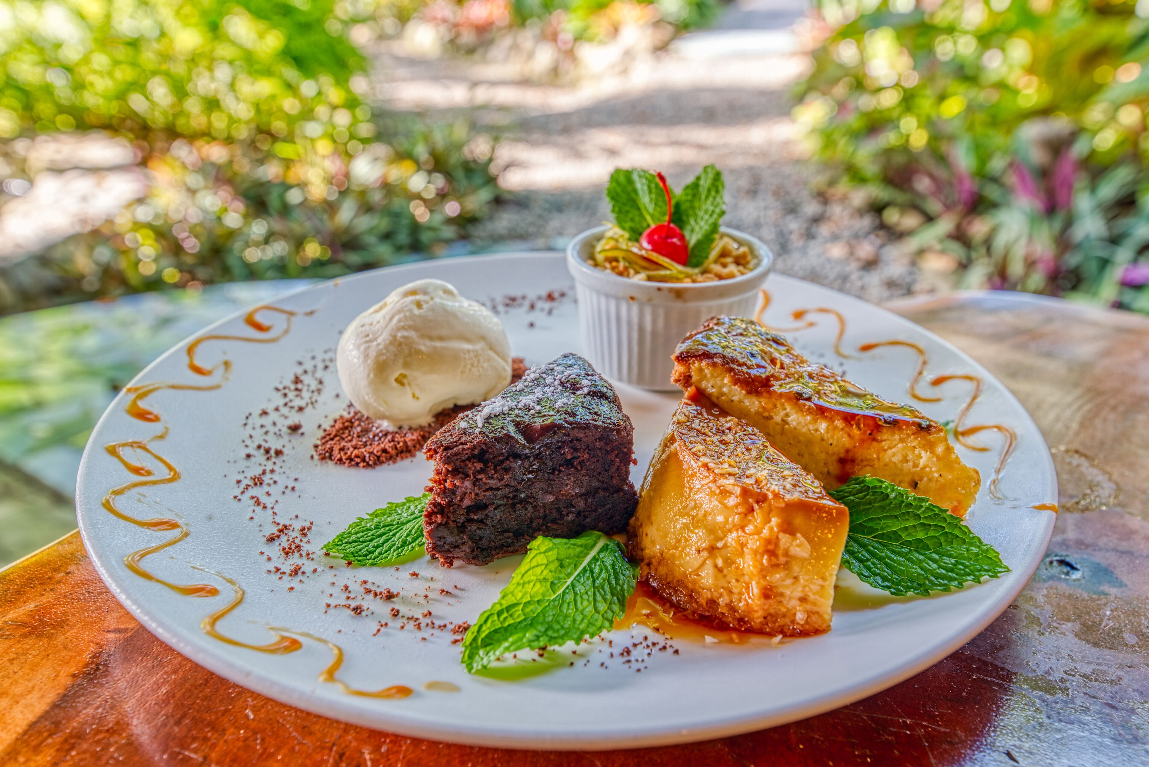 Eine Dessertplatte mit Eis, Schokoladenkuchen und Karamellflan, verziert mit Minzblättern und einer Kirsche, auf einem weißen Teller mit dekorativer Kante.
