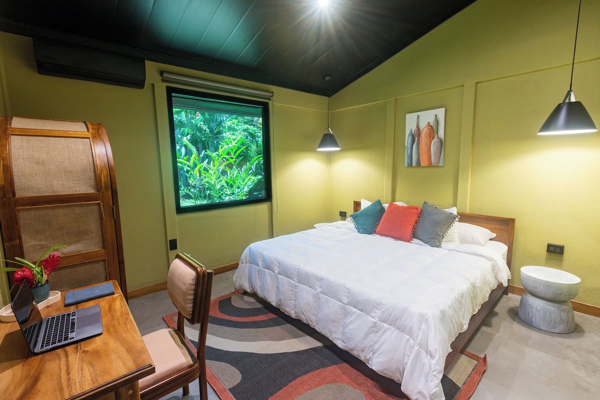 Une chambre avec un lit, un bureau avec un ordinateur portable, une chaise, une fenêtre donnant sur un espace verdoyant et deux lampes suspendues.