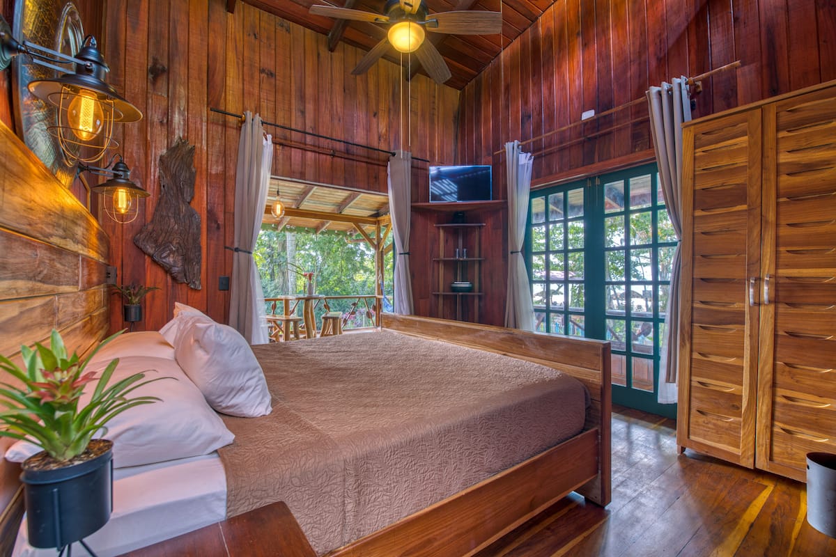Une chambre dans une cabane en bois, équipée d’un lit, d’un ventilateur de plafond, d’une télévision et de grandes fenêtres.