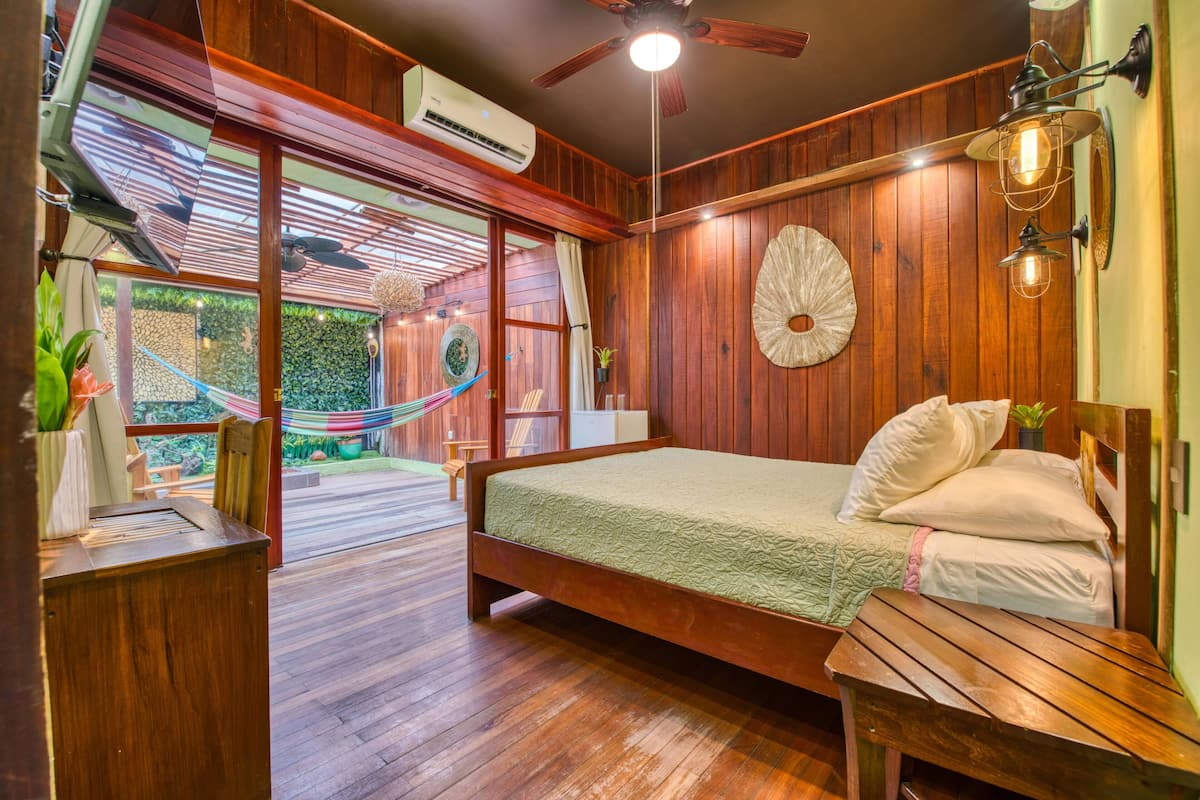 Une chambre avec un plancher en bois, comprenant un lit, un banc, un hamac et donnant sur un jardin.