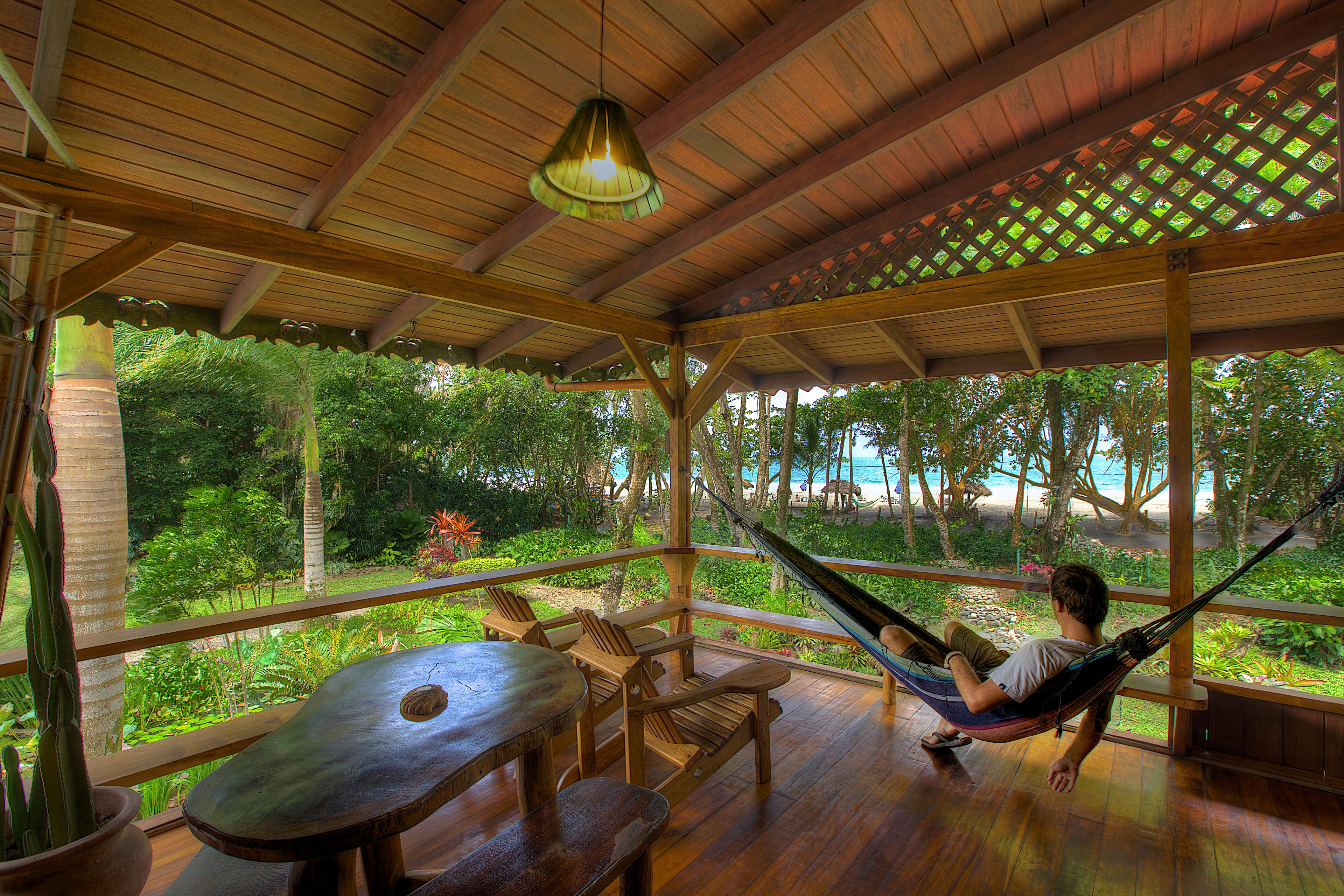 Una terraza de madera con una hamaca, una mesa y sillas, con vistas a un jardín exuberante y una playa.