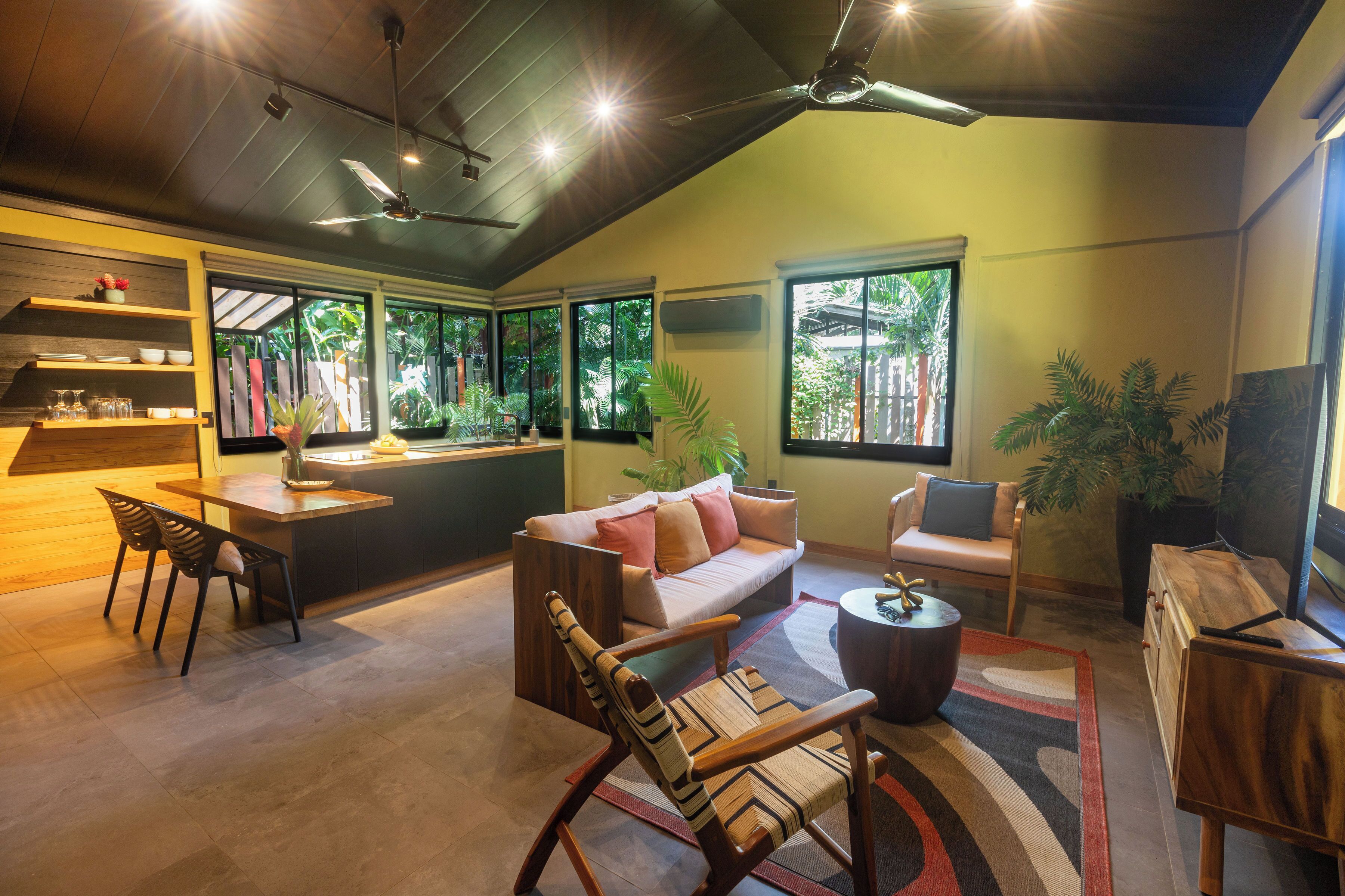A spacious living area with a sofa, chairs, a coffee table, and a dining table. There are large windows providing natural light and views of greenery outside.