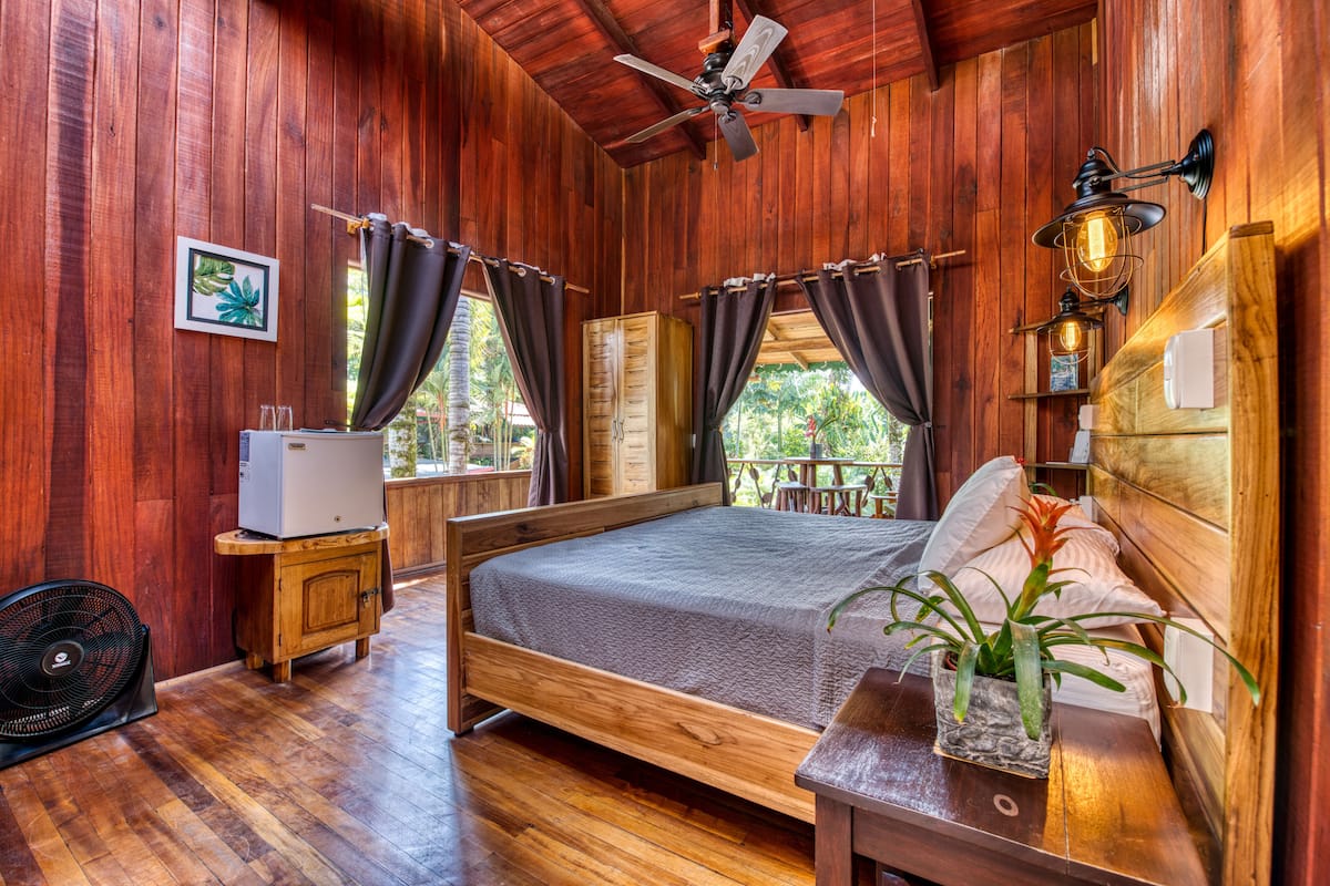 Une pièce en bois avec un lit, une petite armoire, un ventilateur et un ventilateur de plafond.