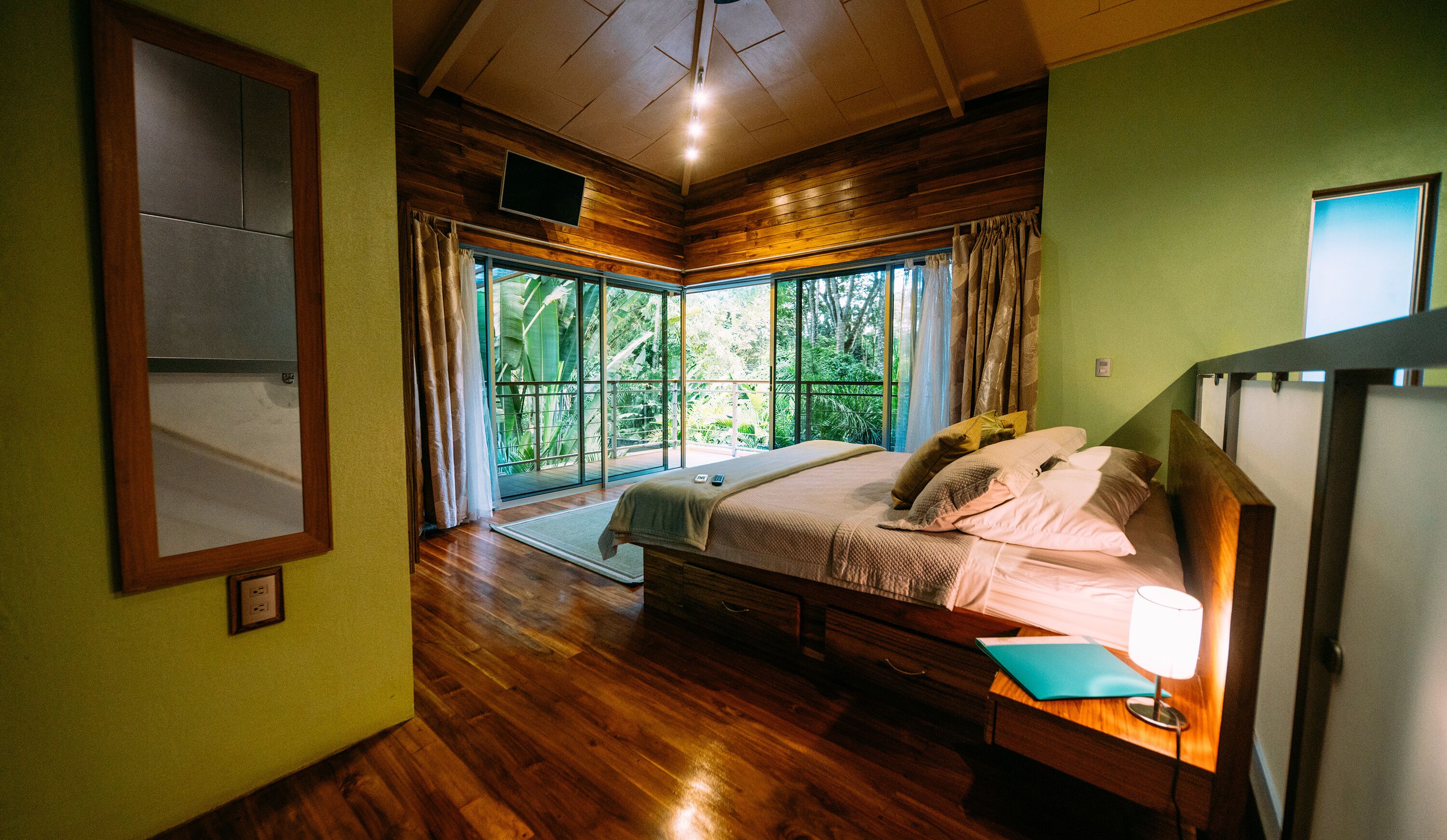 A bedroom with a bed, a bedside table, a window with curtains, and a view of greenery outside.