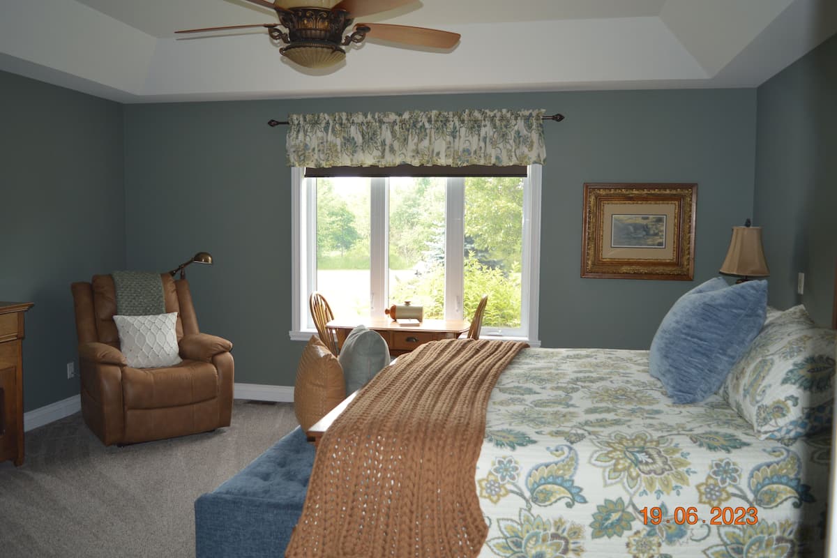 A bedroom with a large bed, a chair, a desk, and a window with a view of trees.
