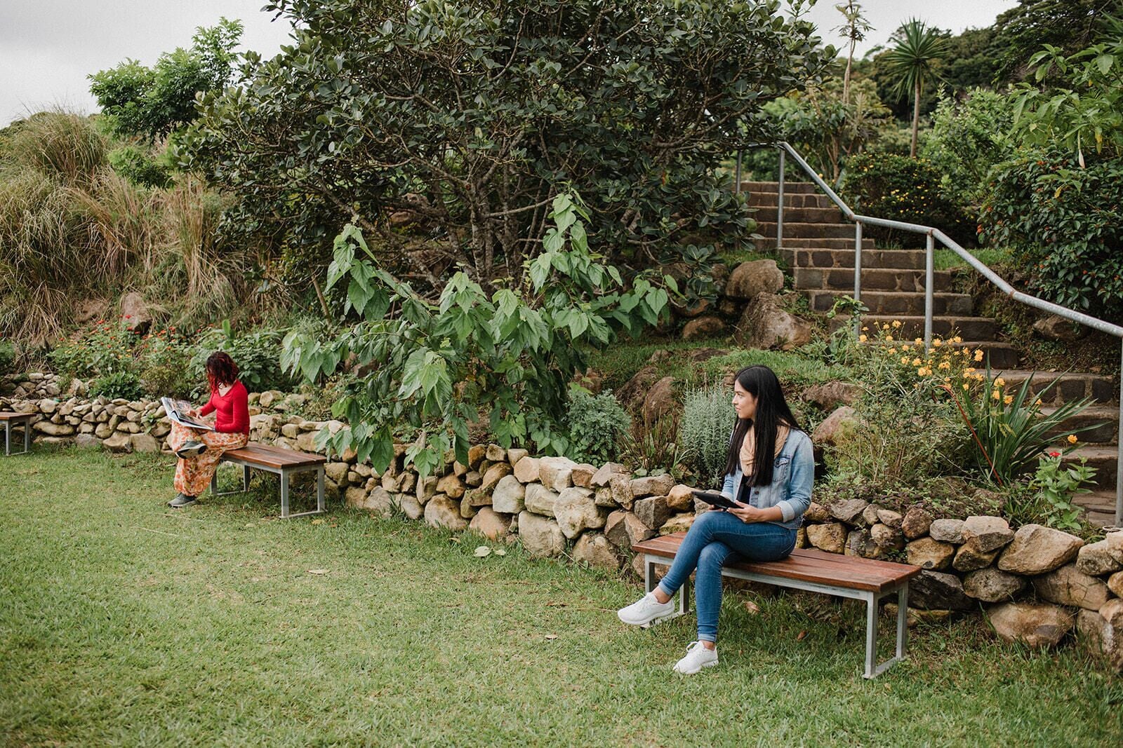 Two people sitting on benches in a garden with a stone wall and a staircase in the background.
