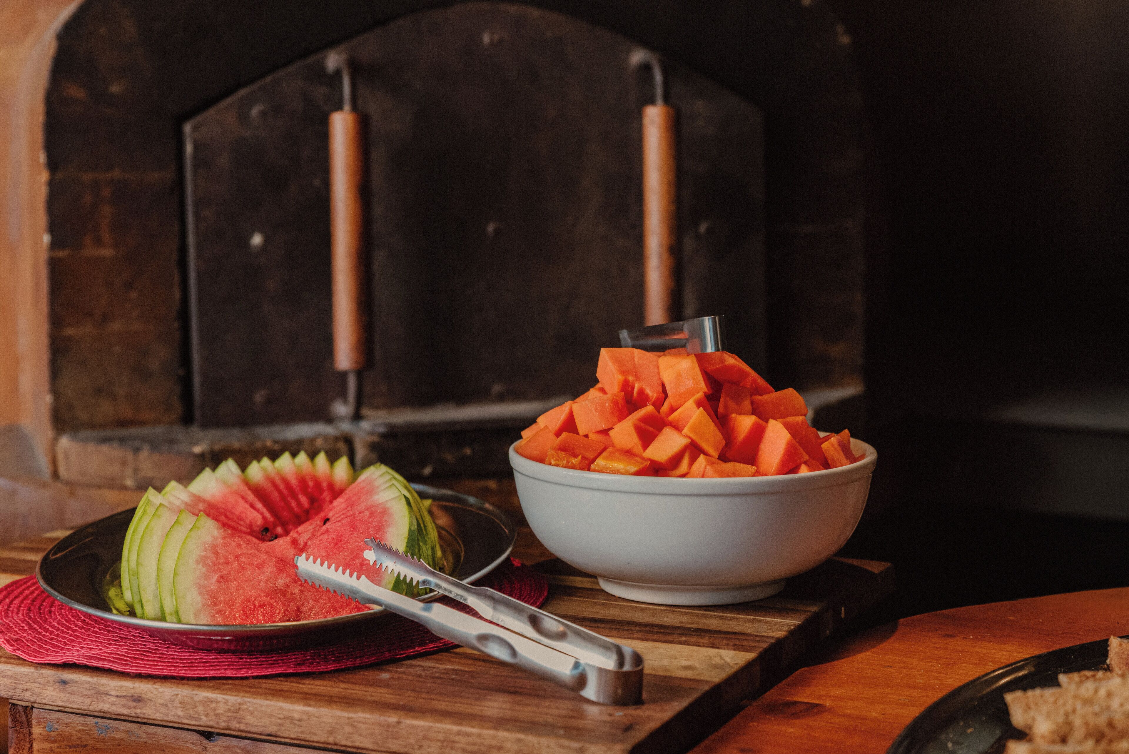A wooden table with a bowl of cubed fruit and a watermelon slice on a red placemat.