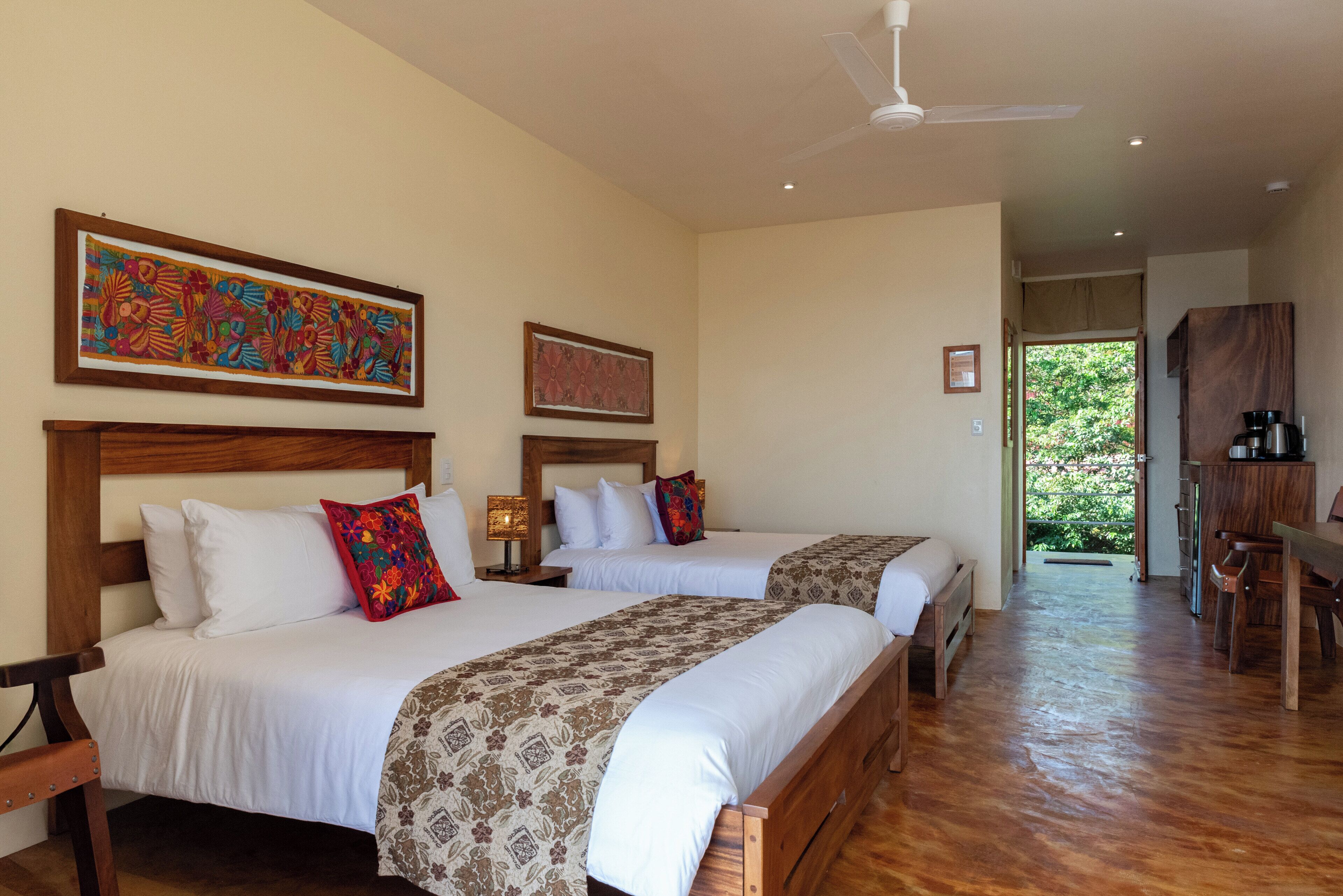 A hotel room with two beds, a ceiling fan, artwork on the walls, and a view of the outdoors.