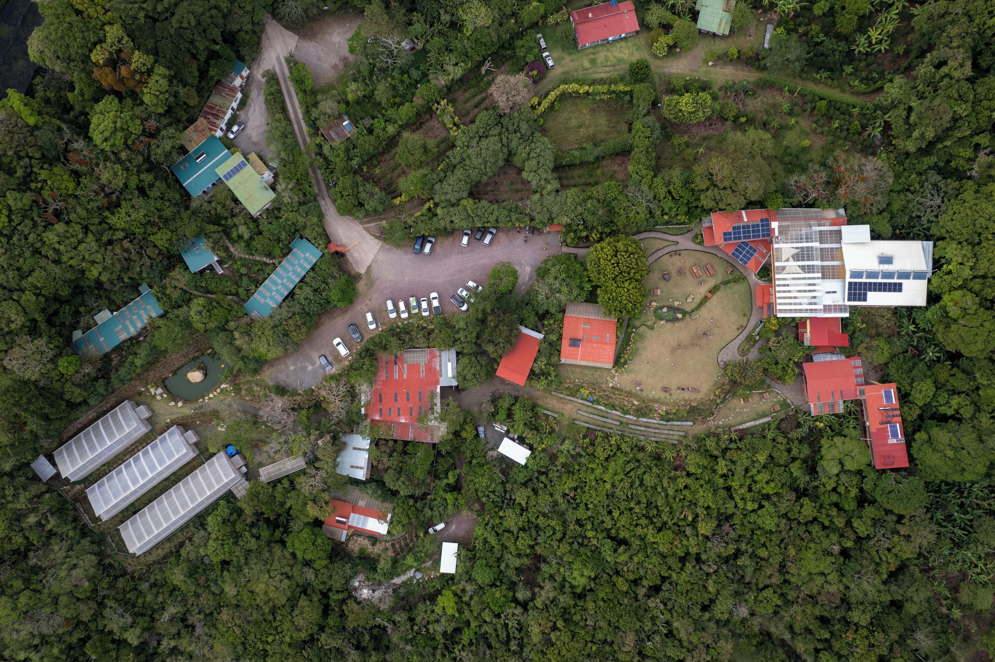 An aerial view of a property with multiple buildings, including a large structure with solar panels, surrounded by dense greenery.