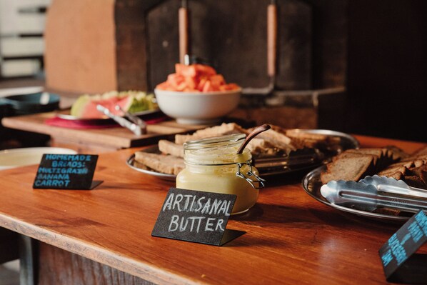 A wooden table with a sign for homemade bread and artisanal butter, a jar of butter, tongs, and a bowl of sliced fruit.