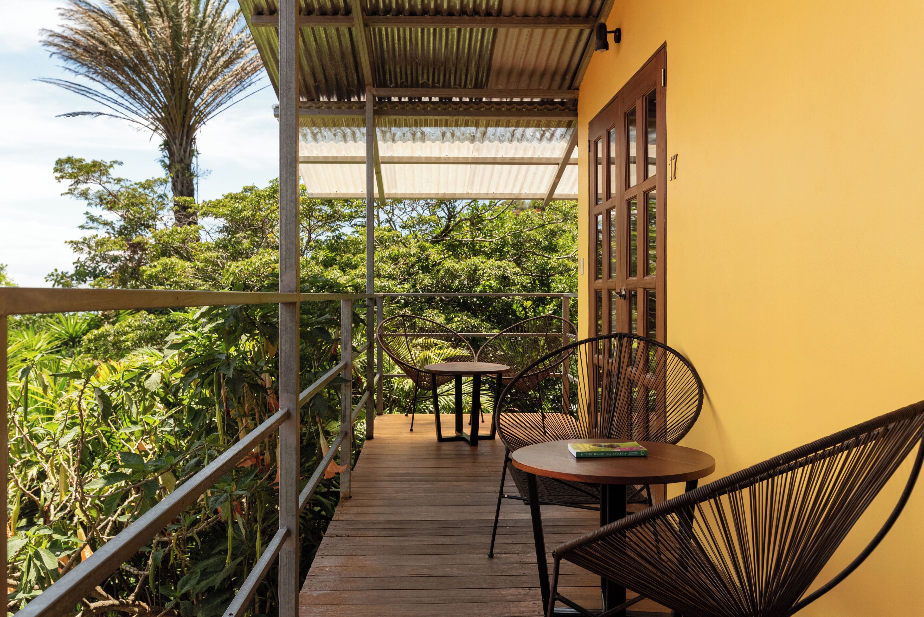 A wooden deck with a table and chairs, surrounded by lush greenery and a yellow wall.