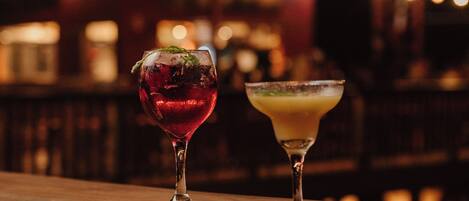 Two cocktails on a bar counter with blurred lights in the background.