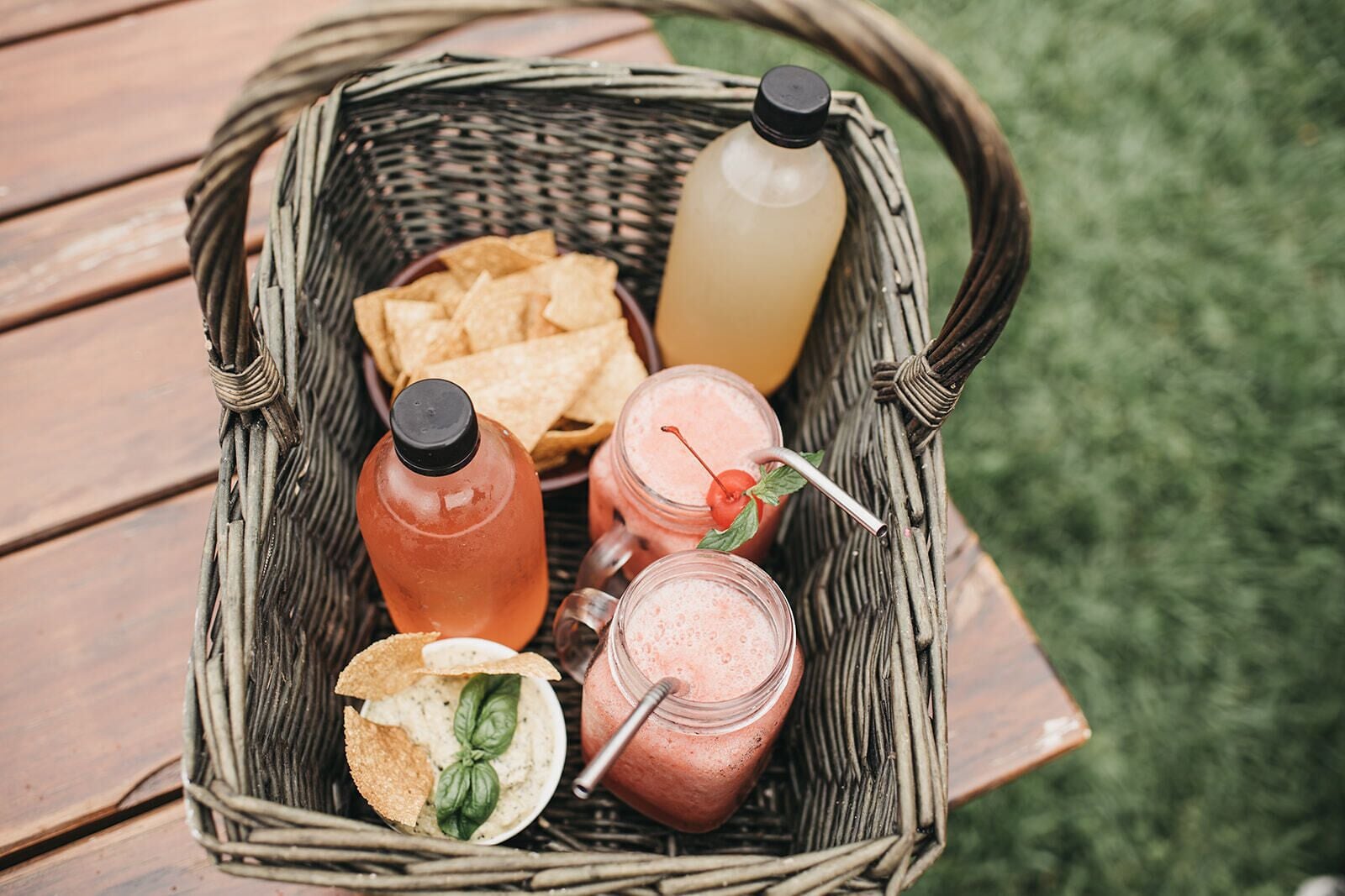 Cesta de vime com uma garrafa de chá gelado, uma garrafa de limonada, um smoothie com cereja, uma tigela de acompanhamento com batatas fritas e uma tortilla chip.