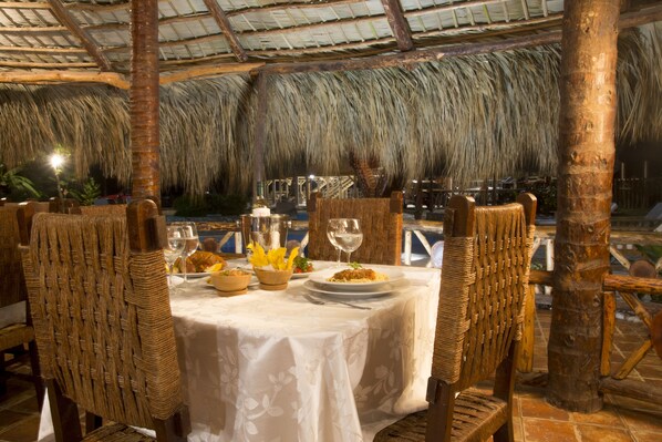 An outdoor dining area with thatched roof, wicker chairs, and a table set for two with a plate of food and wine glasses. - Hotel SeaBreeze (Paraiso)