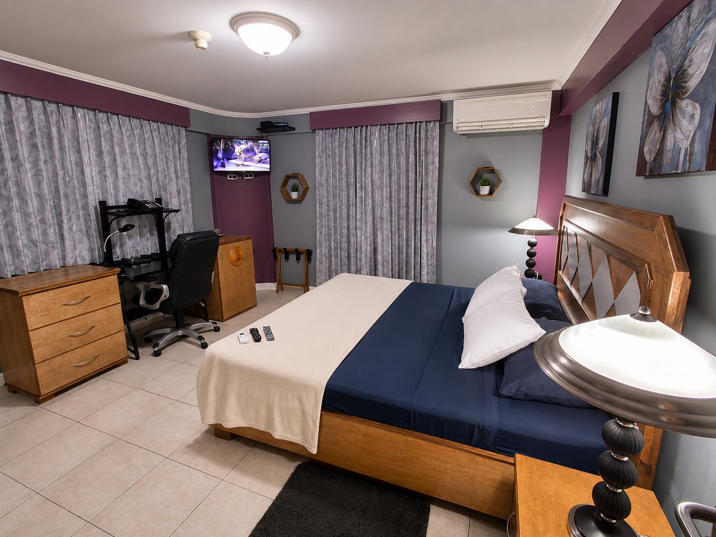 A hotel room with a bed, desk, chair, TV, and a small table with a lamp.