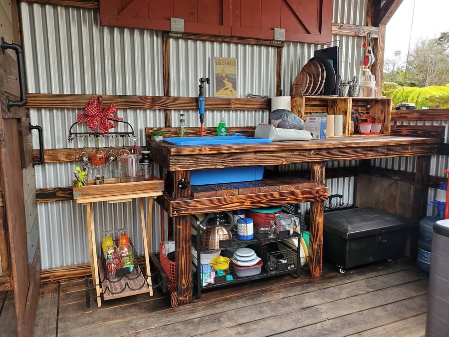 A rustic outdoor kitchen area with a wooden table, shelves, and various kitchen items.