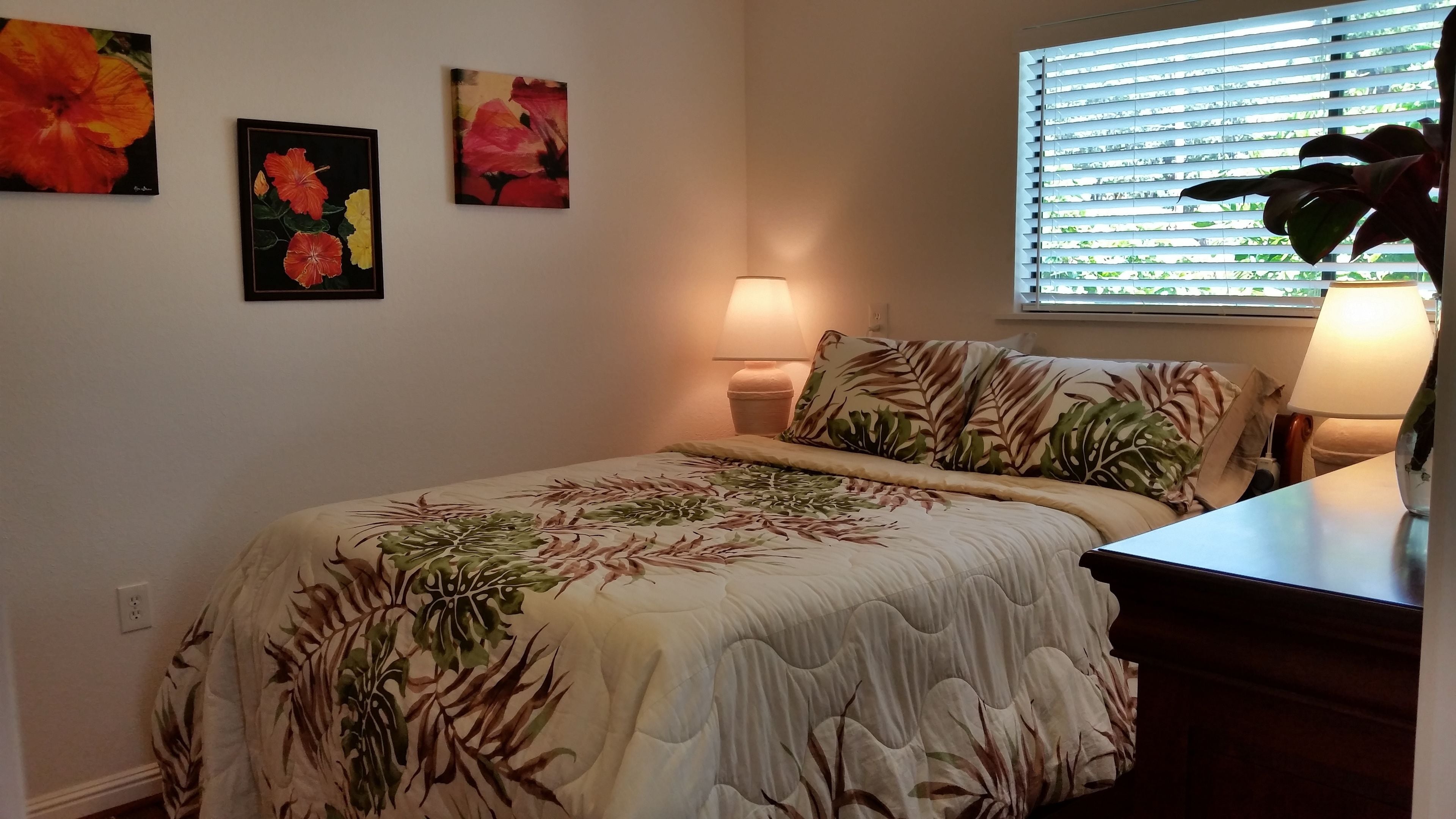Quarto com edredom estampado com flores, abajur e janela com persianas.