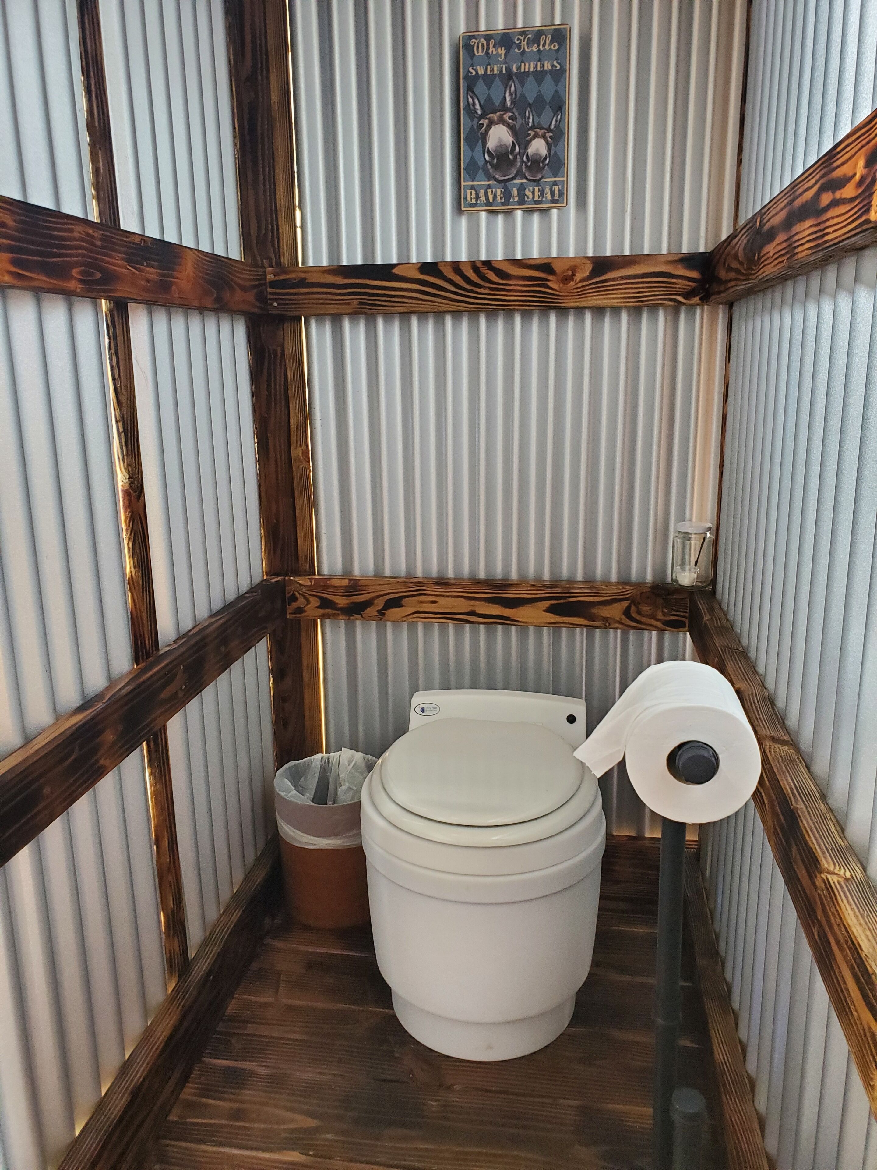A compact, wooden-framed bathroom with a corrugated metal wall, a toilet, and a roll of toilet paper.