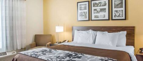 A hotel room with a bed, a chair, a bedside table, a lamp, and three framed pictures on the wall.