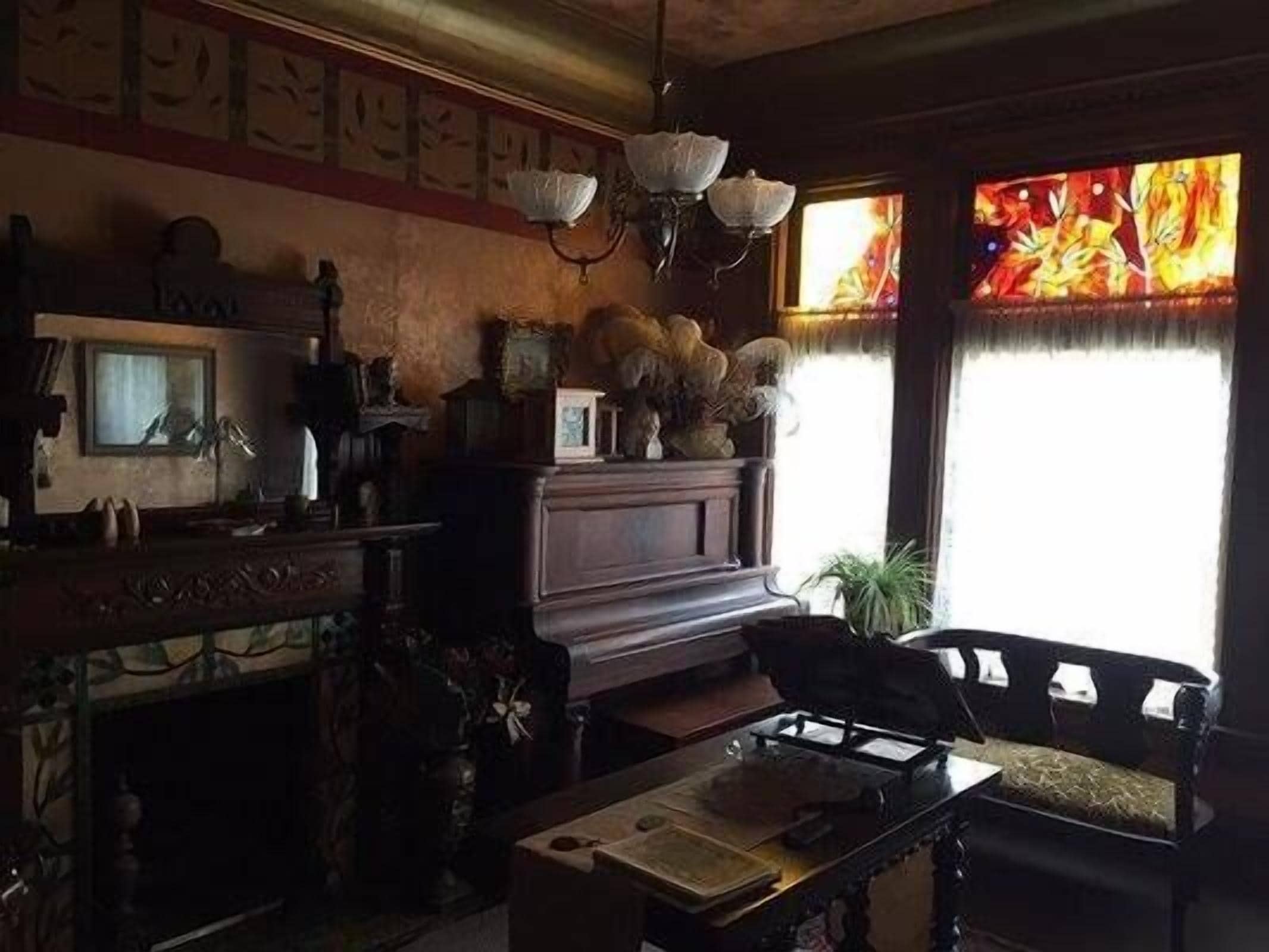 A dimly lit room with a piano, a wooden cabinet, and a chair. There is a window with a stained glass design.