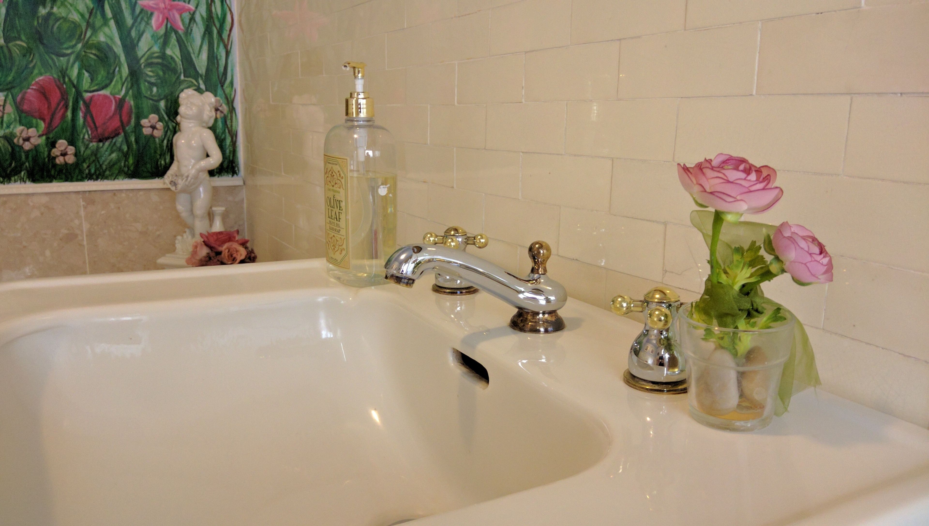 Une salle de bain avec un double lavabo, un robinet doré, une bouteille d’huile d’olive et un vase rempli de fleurs roses.