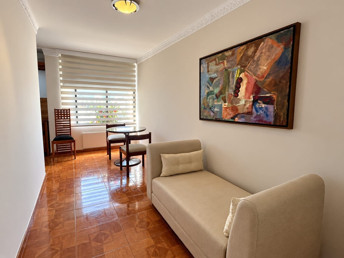 Uma sala de estar com sofá, mesa de jantar e cadeiras, um quadro na parede e uma janela com persianas.
