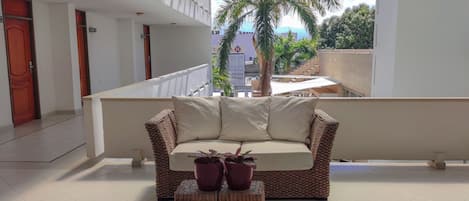 Un balcón con un sofá de mimbre, dos plantas en macetas y vistas a palmeras y edificios.