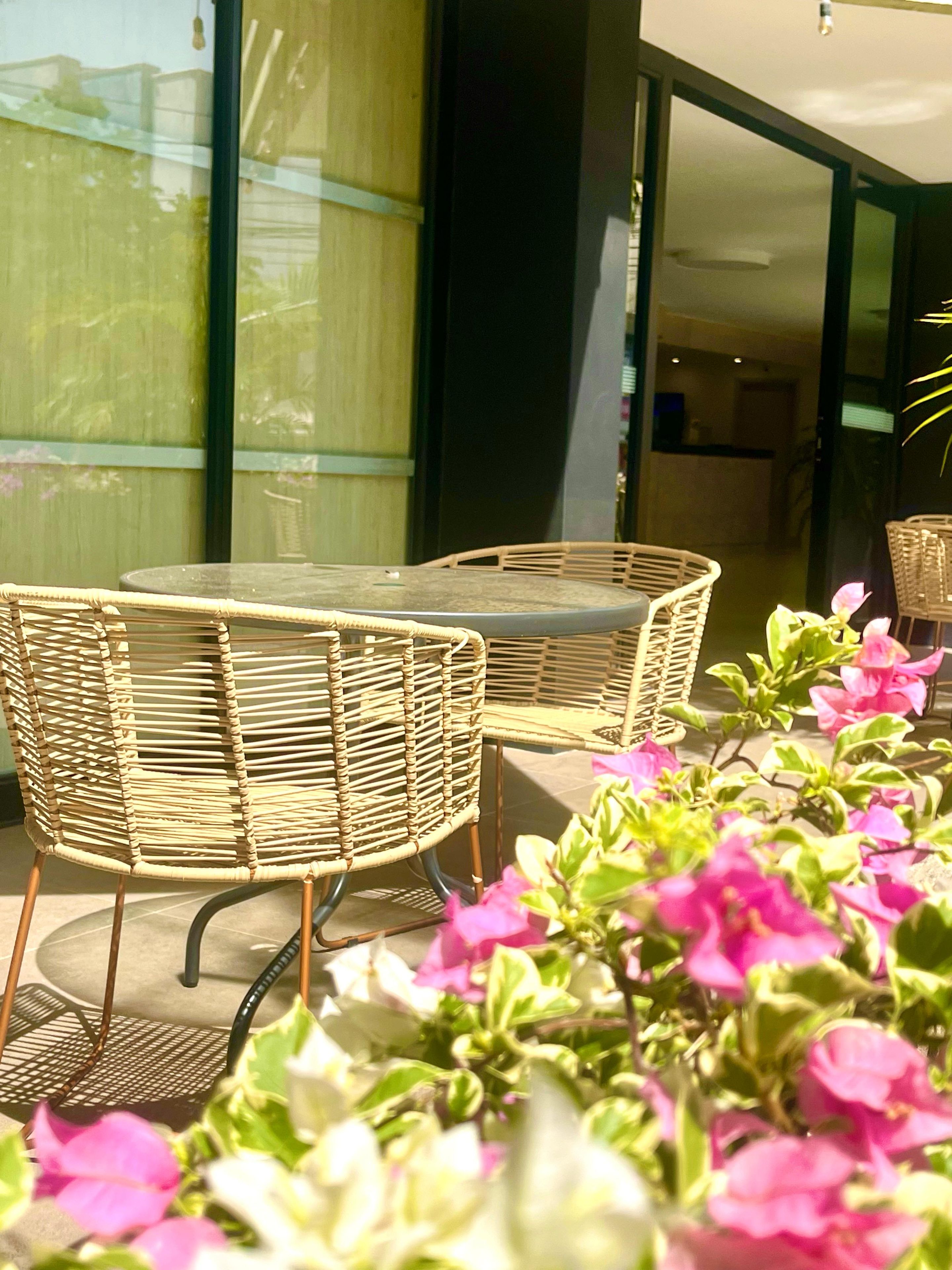 Un espace extérieur aménagé avec des meubles en osier, comprenant une table en verre et des chaises, le tout entouré de plantes en pot aux fleurs roses.