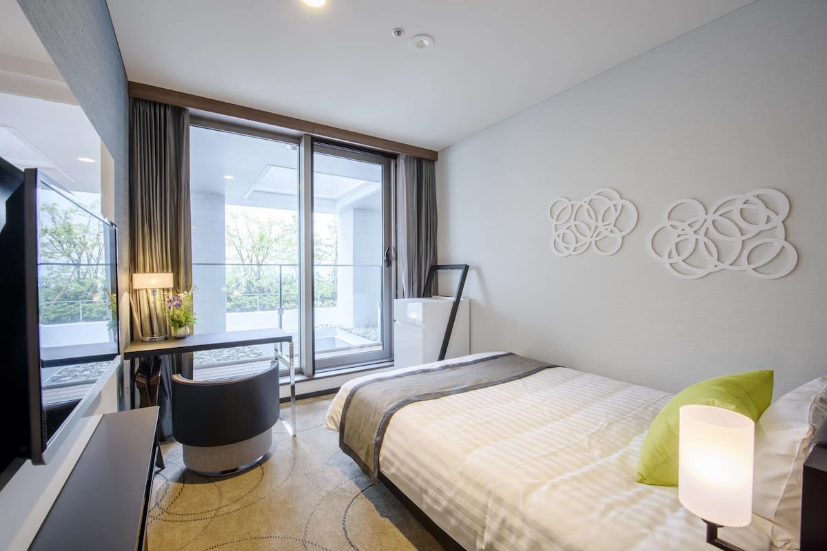 A modern hotel room with a large bed, a desk, and a view of the outdoors through a sliding door.