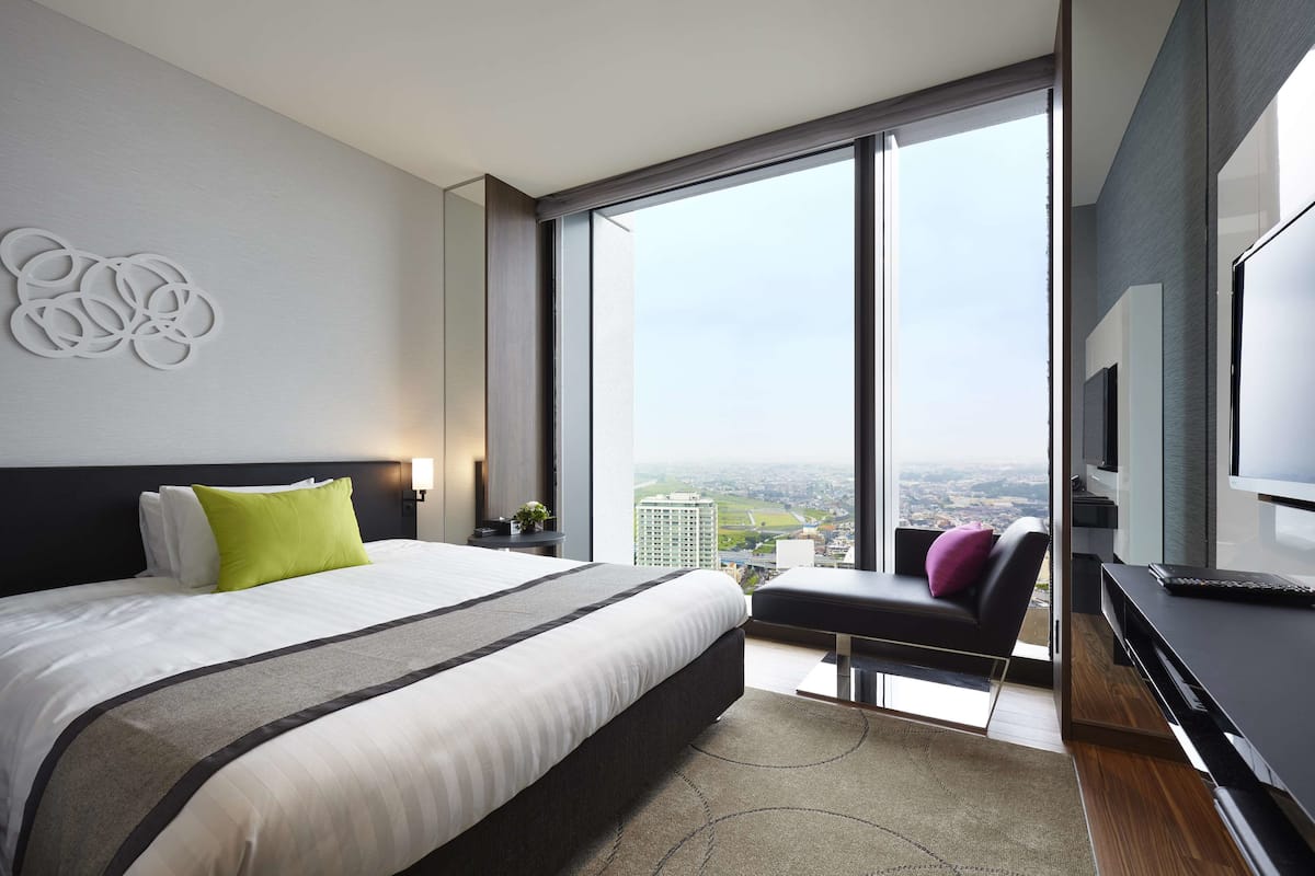 A modern hotel room with a large bed, a seating area, a TV, and a city view.
