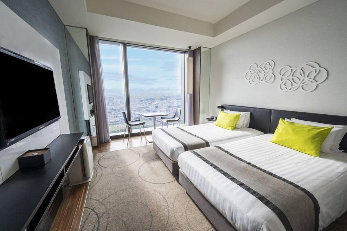 A hotel room with two beds, a TV, a desk, and a view of the city.