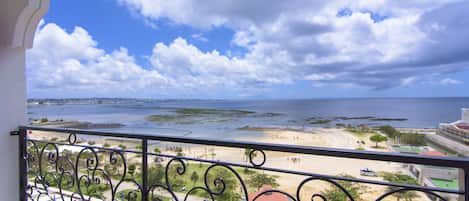 A balcony with a view of a beach and ocean, featuring a metal railing with decorative scrollwork.