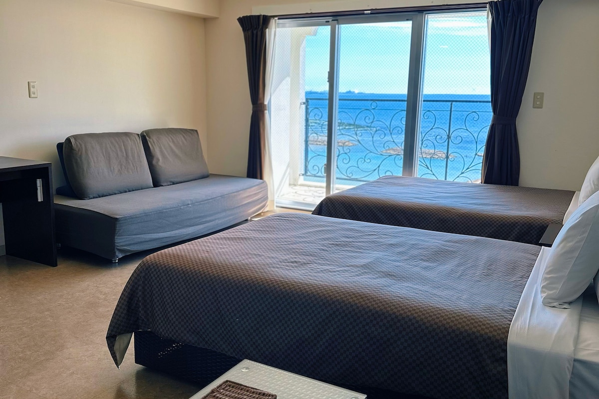 A hotel room with two beds, a sofa, a small table, and a view of the sea.
