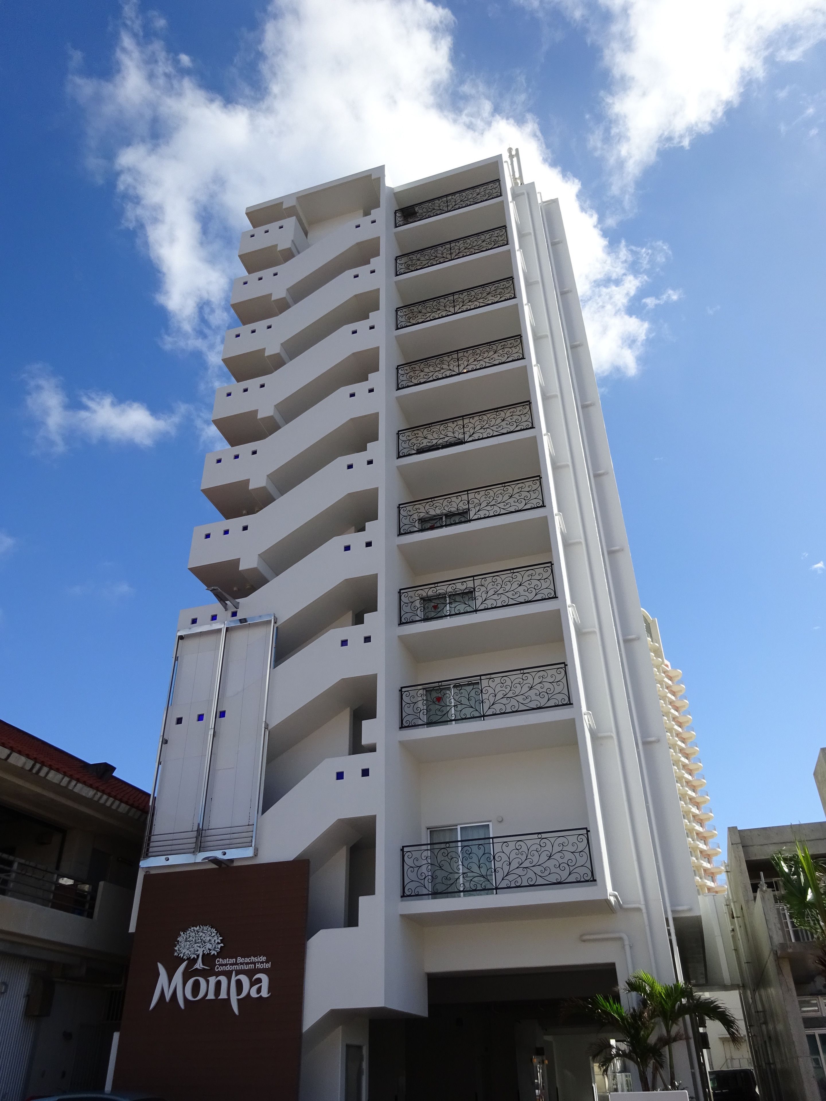 Um edifício alto, com várias andares e varandas, com uma placa indicando 'Monpa Chatan Beachside Condominium Hotel' e um céu azul claro ao fundo.