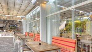 An outdoor café with wooden tables and striped cushioned seating, a stone wall with a neon sign, and large windows offering a view of the outdoors. - D'primahotel Tangerang (Tangerang)