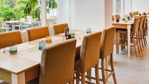 A spacious dining area with long wooden tables and chairs, large windows offering a view of greenery outside, and pendant lighting. - D'primahotel Tangerang (Tangerang)