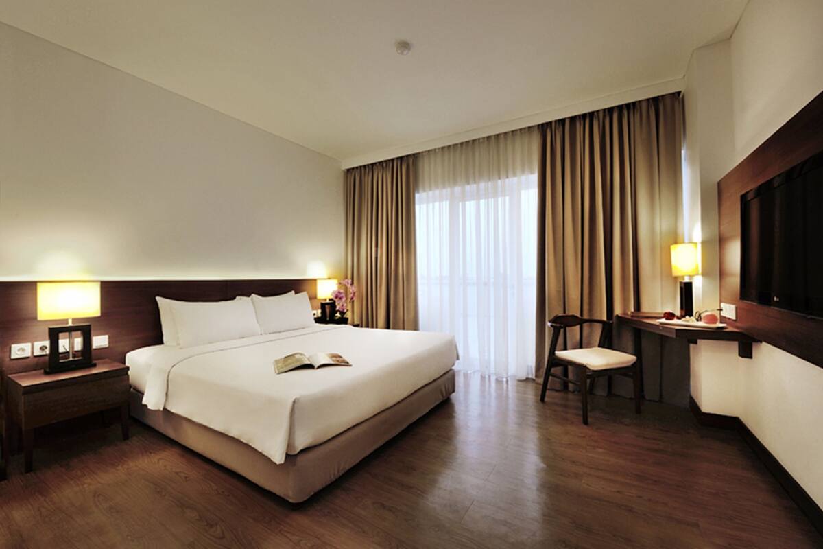 Quarto de hotel com uma cama grande, luminárias de cabeceira, uma mesa com cadeira e uma janela ampla com cortinas.