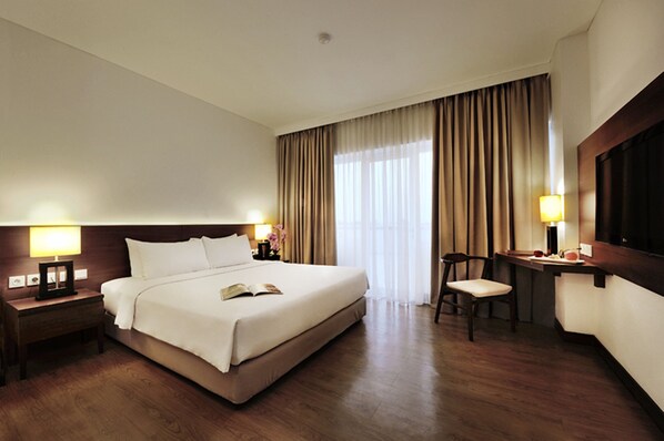 A hotel room with a large bed, bedside lamps, a desk with a chair, and a large window with curtains. - D'primahotel Tangerang (Tangerang)