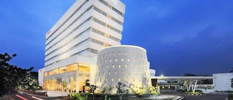 Un hotel moderno con un llamativo letrero que dice 'prima hotel', un edificio circular con ventanas uniformemente distribuidas, y una zona ajardinada bien cuidada con árboles.