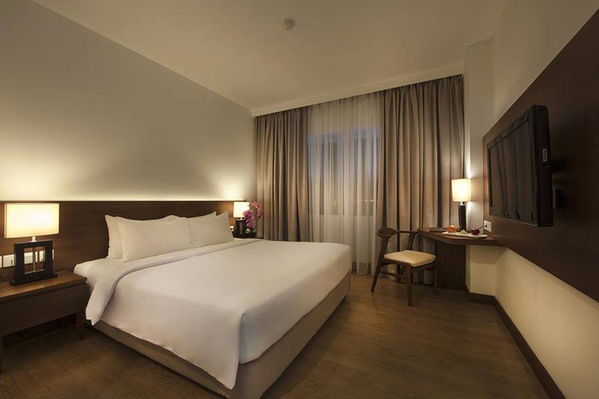 Quarto de hotel com uma cama grande, luminárias de cabeceira, uma escrivaninha com cadeira e uma TV fixada na parede.