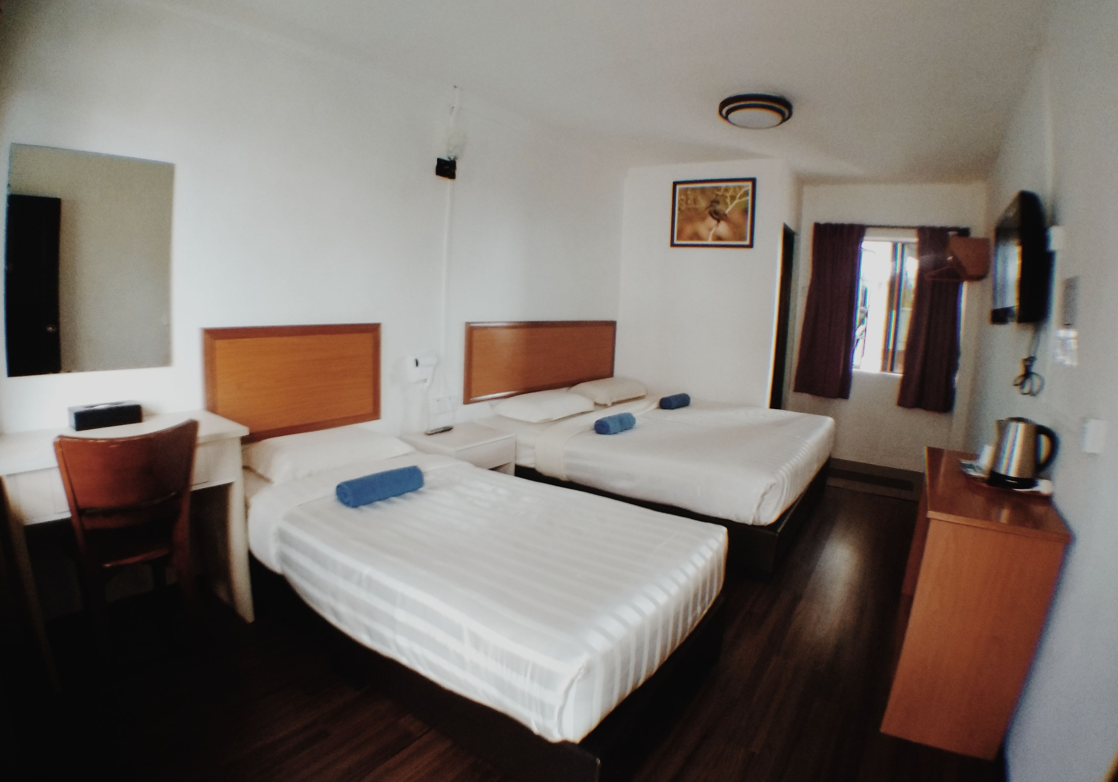 A hotel room with two single beds, a desk with a chair, a small table with a kettle, and a mirror on the wall.