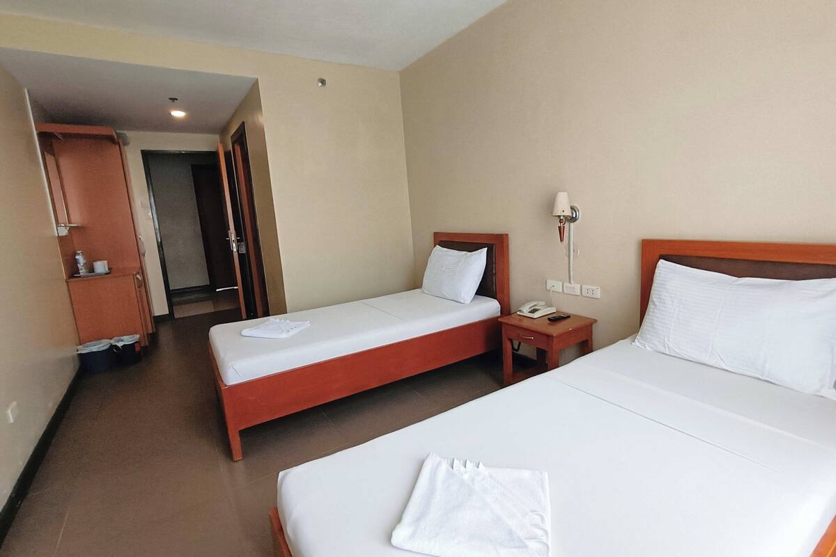 A hotel room with two single beds, a wooden wardrobe, a bedside table with a lamp, and a door leading to another room.