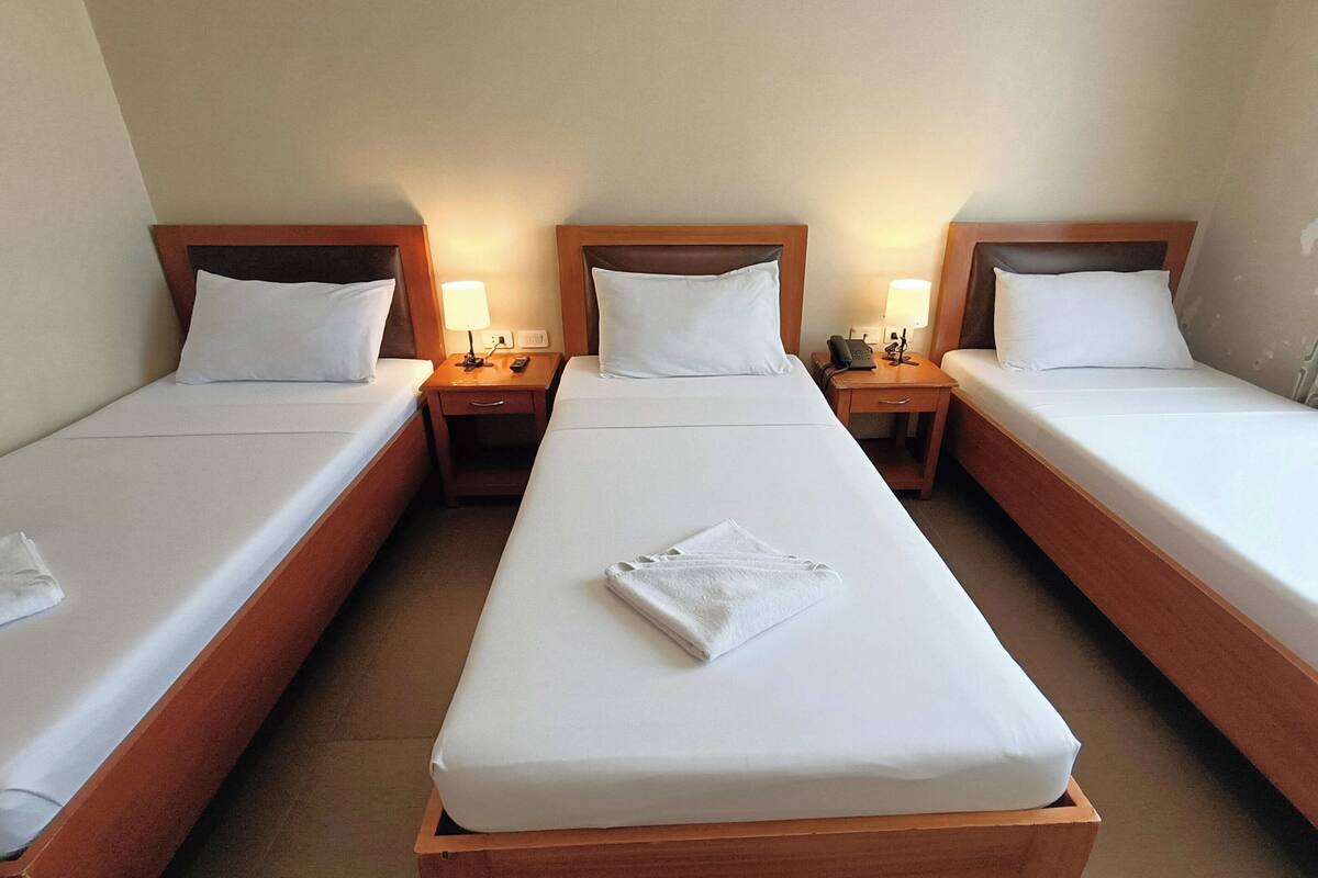 A hotel room with three single beds, each with a wooden headboard, white linens, and bedside lamps.